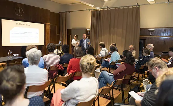Deelnemers in een workshop kijken zittend naar slides die gepresteneerd worden door twee sprekers